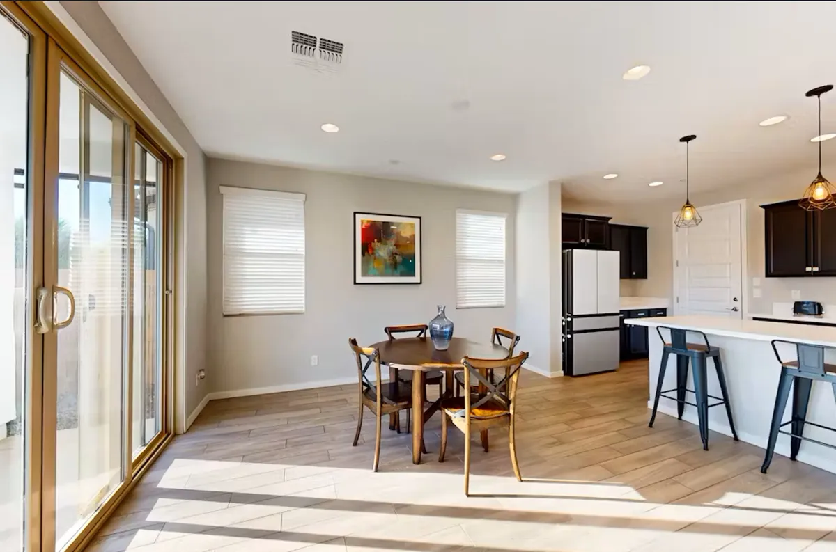 A bright, airy view centered on a dining table adjacent to a kitchen island, with large windows flooding the area with sunlight. The scene suggests a relaxed setting for family meals, casual gatherings, and conversations, enhanced by thoughtful artwork on the wall.