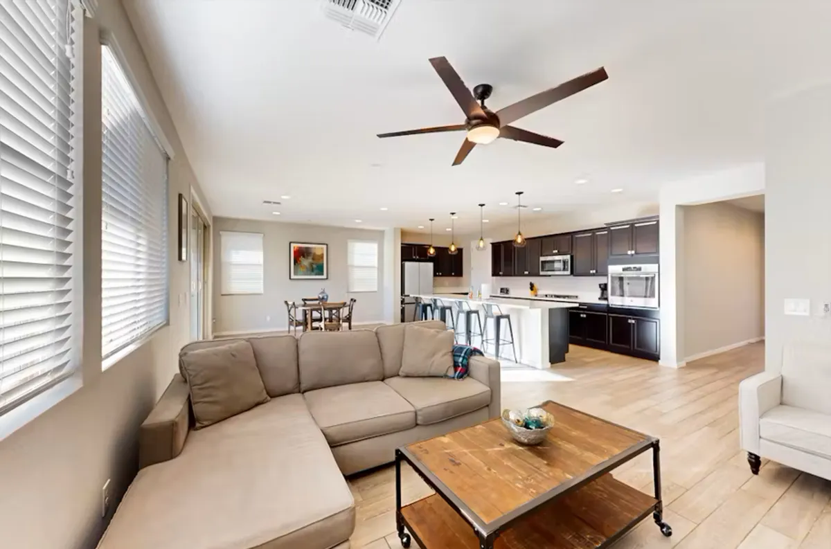A wide, inviting shot of an open-concept living area featuring a sectional sofa, wooden coffee table, kitchen island, and dining table. Natural light pours in through large windows, illuminating the cohesive space where guests can relax, dine, or prepare meals together.