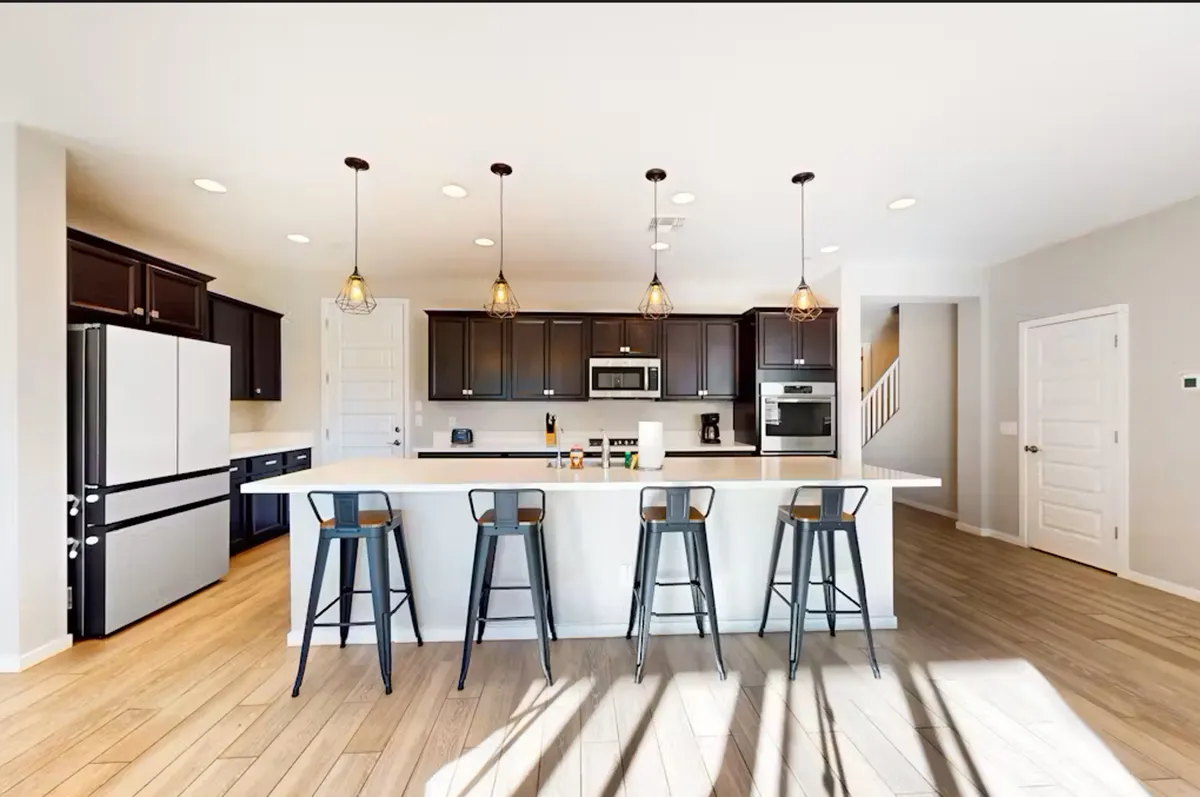 This image showcases a stylish kitchen with an island countertop, pendant lights, bar stools, dark cabinetry, and modern appliances. The space is well-lit and designed for gathering, making it ideal for entertaining or casual group meals.