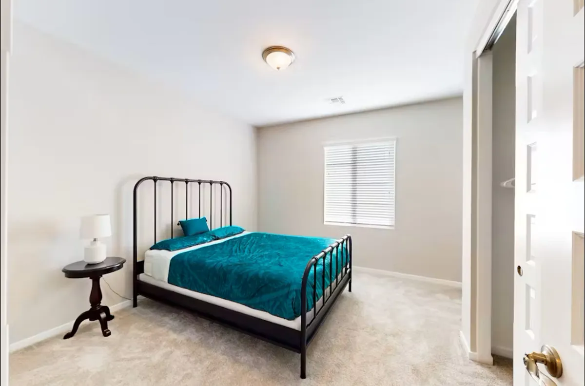 A tranquil bedroom with a bed dressed in teal bedding, accented by a nightstand with a lamp and a window with blinds. The space feels private and inviting, ideal for relaxation and reading.