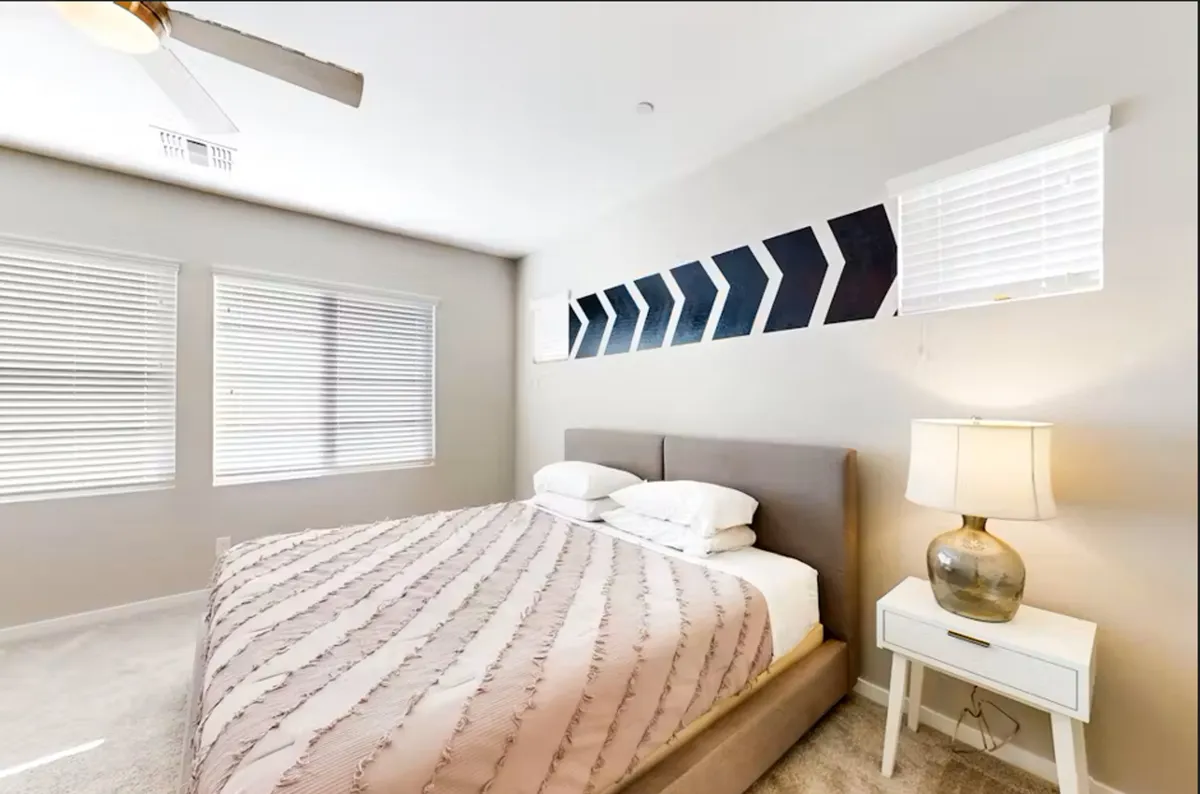 A serene bedroom featuring a neatly made bed, decorative wall design, a cozy lamp on the nightstand, and windows that flood the room with natural light. The soft textures and calming color scheme create a peaceful atmosphere.