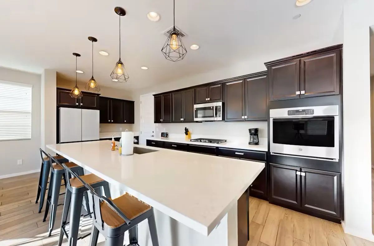 A large, contemporary kitchen island is the focal point, surrounded by pendant lighting, dark cabinetry, and stainless steel appliances. The scene is lively and functional, showing a space fit for hosting friends, family game nights, or casual brunches.