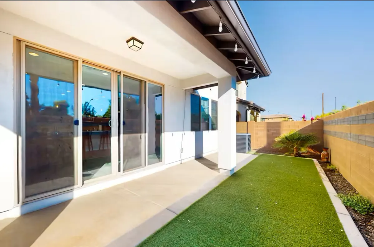 This exterior image showcases a backyard with artificial grass, decorative palm tree, sliding glass doors, and welcoming outdoor lighting fixtures. The scene suggests a private, serene outdoor space for relaxation or hosting small gatherings.