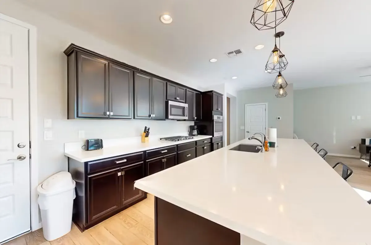 This image features a bright, welcoming kitchen with a spacious island countertop, dark cabinets, and modern hanging light fixtures. The scene suggests a comfortable space where families can gather after a fun day out, cook together, or entertain friends.
