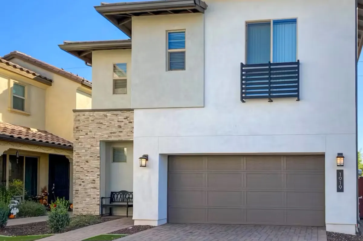 The image showcases the home's modern architectural design with a striking stone facade, an inviting balcony with elegant railing, and a spacious two-car garage. The front yard is ideal for gatherings or outdoor relaxation, bathed in warm Arizona sunlight, with ample space for barbecues or enjoying sunsets from the balcony.